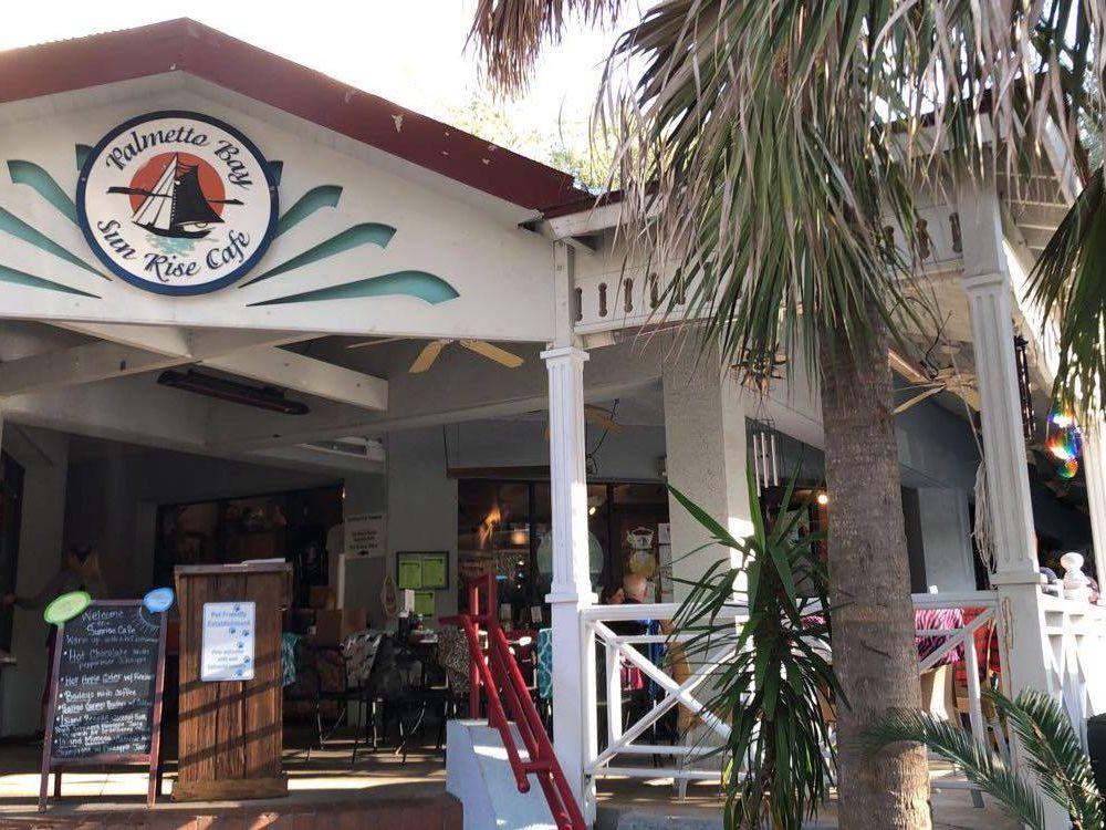 The entrance of Palmetto Bay Sunrise Cafe, a cozy local breakfast spot surrounded by palm trees and coastal charm. Guests gather on the shaded patio to enjoy coffee and classic Southern breakfast dishes. This popular Hilton Head cafe is known for relaxed island vibes and fresh morning favorites.
