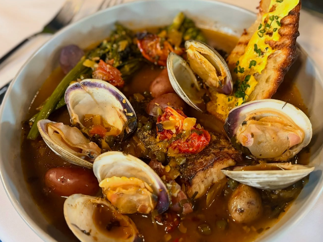 A hearty seafood stew featuring clams, fish, potatoes, broccolini, and cherry tomatoes in a rich broth, served with a grilled slice of bread topped with saffron aioli and chopped chives in a wide bowl.