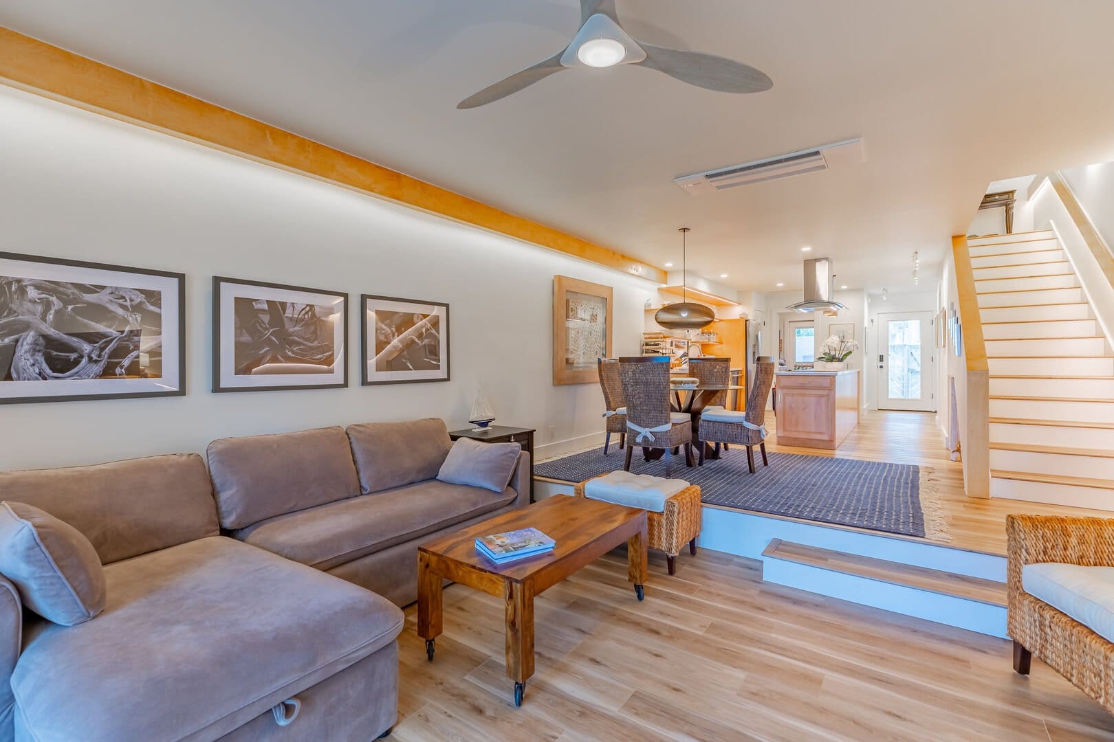 Open-concept living space with a neutral-toned sectional sofa, wooden coffee table, and woven accent chairs, leading to a dining area and modern kitchen. A staircase with sleek white steps ascends to the second floor, while warm lighting and natural wood elements create a cozy, contemporary feel.