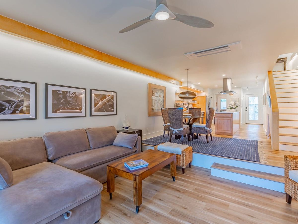 Open-concept living space with a neutral-toned sectional sofa, wooden coffee table, and woven accent chairs, leading to a dining area and modern kitchen. A staircase with sleek white steps ascends to the second floor, while warm lighting and natural wood elements create a cozy, contemporary feel.