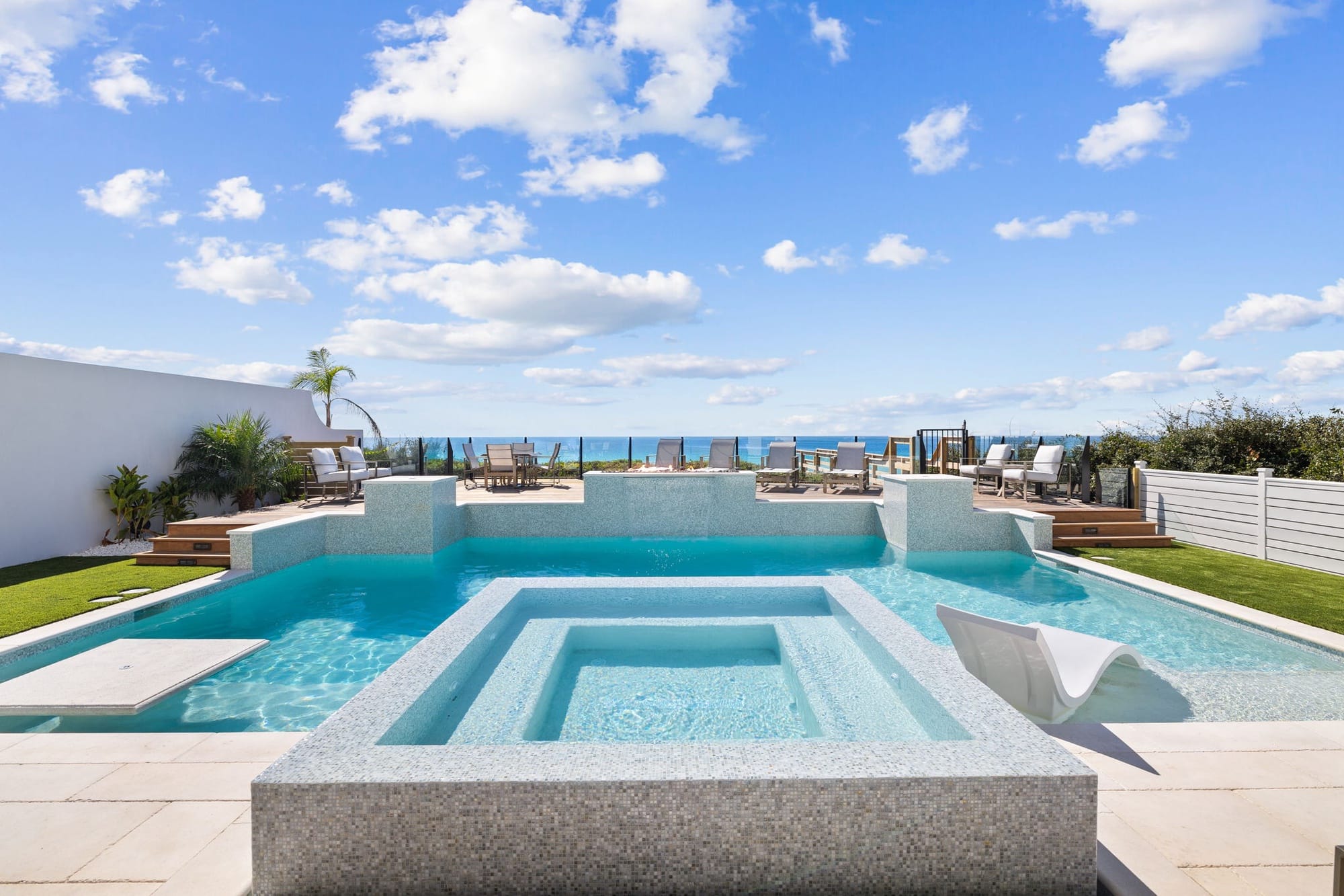 A stunning resort-style pool and spa at Villa Grandeur feature iridescent mosaic tile, a sculptural white sun lounger, and a raised sun deck with lounge chairs overlooking the emerald waters of the Gulf of Mexico. Tropical landscaping, artificial turf, and a glass perimeter fence complete the luxurious outdoor oasis under a bright blue sky. 