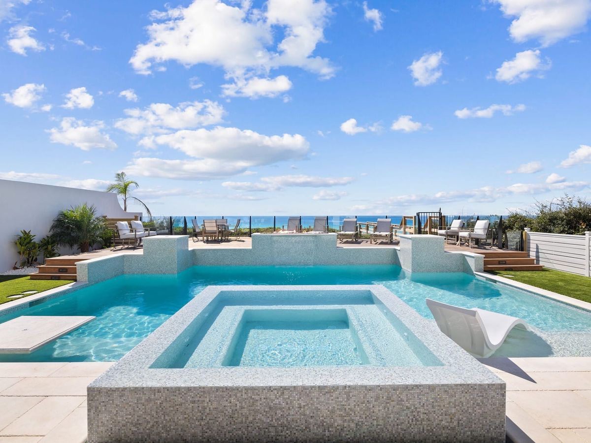 A stunning resort-style pool and spa at Villa Grandeur feature iridescent mosaic tile, a sculptural white sun lounger, and a raised sun deck with lounge chairs overlooking the emerald waters of the Gulf of Mexico. Tropical landscaping, artificial turf, and a glass perimeter fence complete the luxurious outdoor oasis under a bright blue sky.