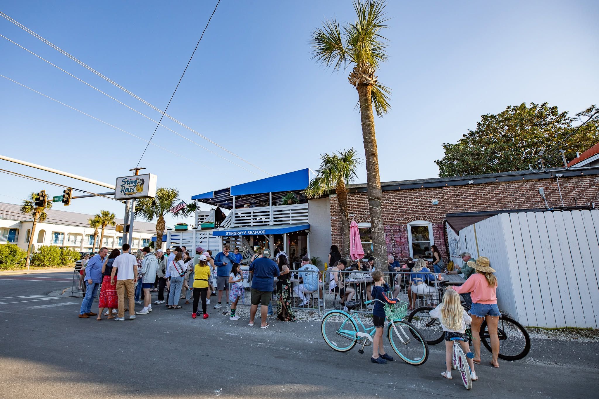 A crowd of people waits outside StingRay's Seafood, a brick beachside restaurant with a blue awning and rooftop deck, as tall palm trees rise against a clear blue sky. Beachgoers with bicycles gather near the street corner beneath the restaurant's sign on a sunny day. 