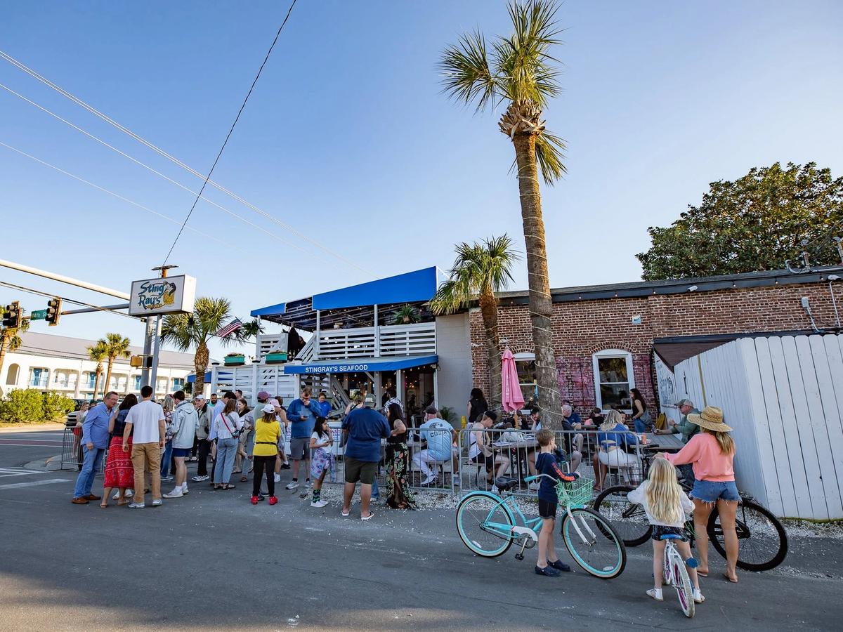 A crowd of people waits outside StingRay's Seafood, a brick beachside restaurant with a blue awning and rooftop deck, as tall palm trees rise against a clear blue sky. Beachgoers with bicycles gather near the street corner beneath the restaurant's sign on a sunny day.