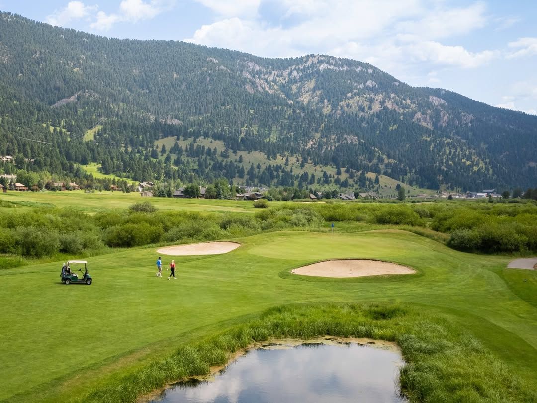 A wide golf course sits under a mountain ridge in Big Sky, with golfers walking near a cart and sand bunkers. It’s a peaceful and scenic place to play a round while surrounded by green fields and forested hills.
