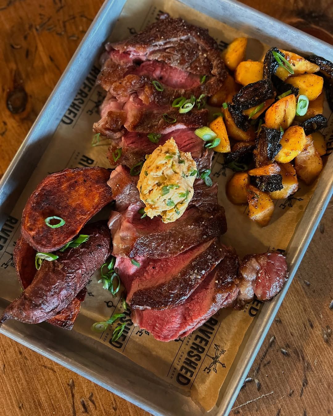Sliced medium-rare steak served on a tray with roasted potatoes and mushrooms at Rivershed in Killington. The dish is topped with herb butter and fresh green onions, creating a rich and savory mountain dining experience. This popular Killington restaurant is known for hearty comfort food after a day on the slopes.