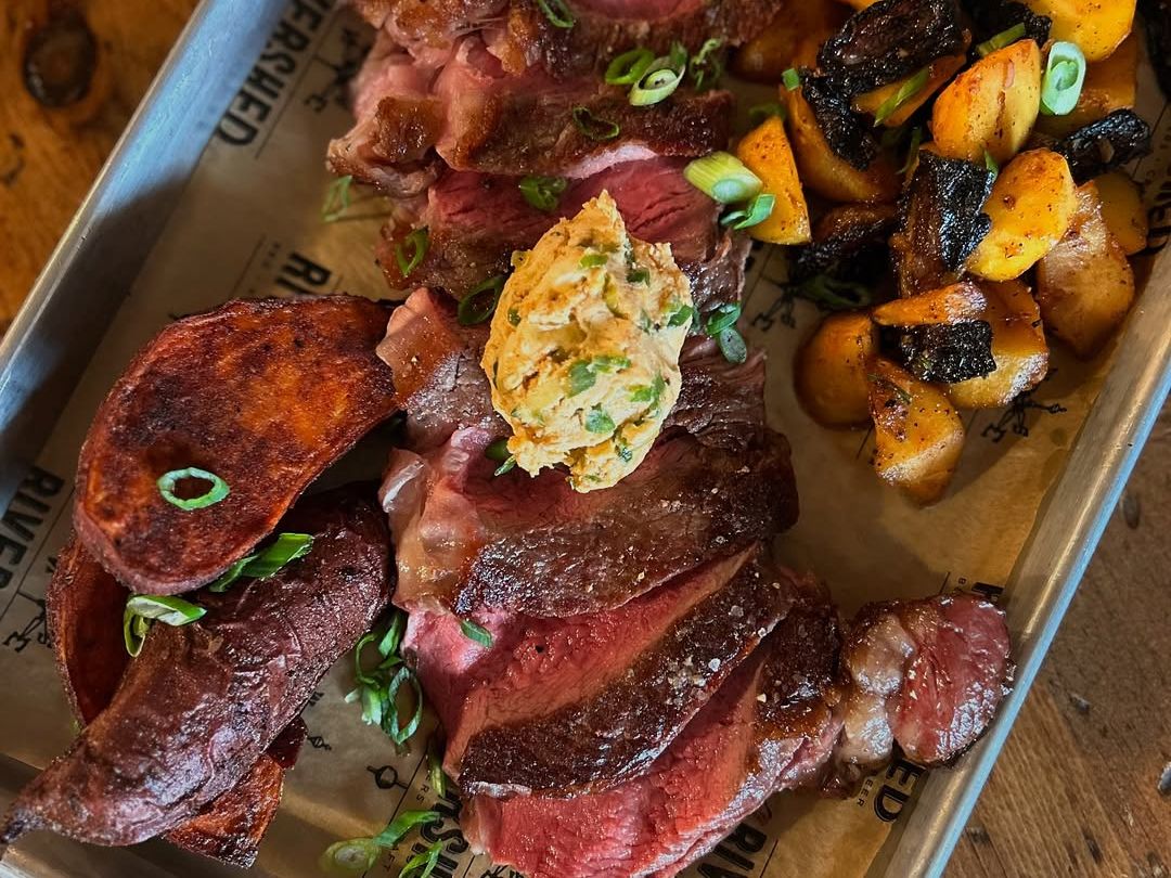 Sliced medium-rare steak served on a tray with roasted potatoes and mushrooms at Rivershed in Killington. The dish is topped with herb butter and fresh green onions, creating a rich and savory mountain dining experience. This popular Killington restaurant is known for hearty comfort food after a day on the slopes.