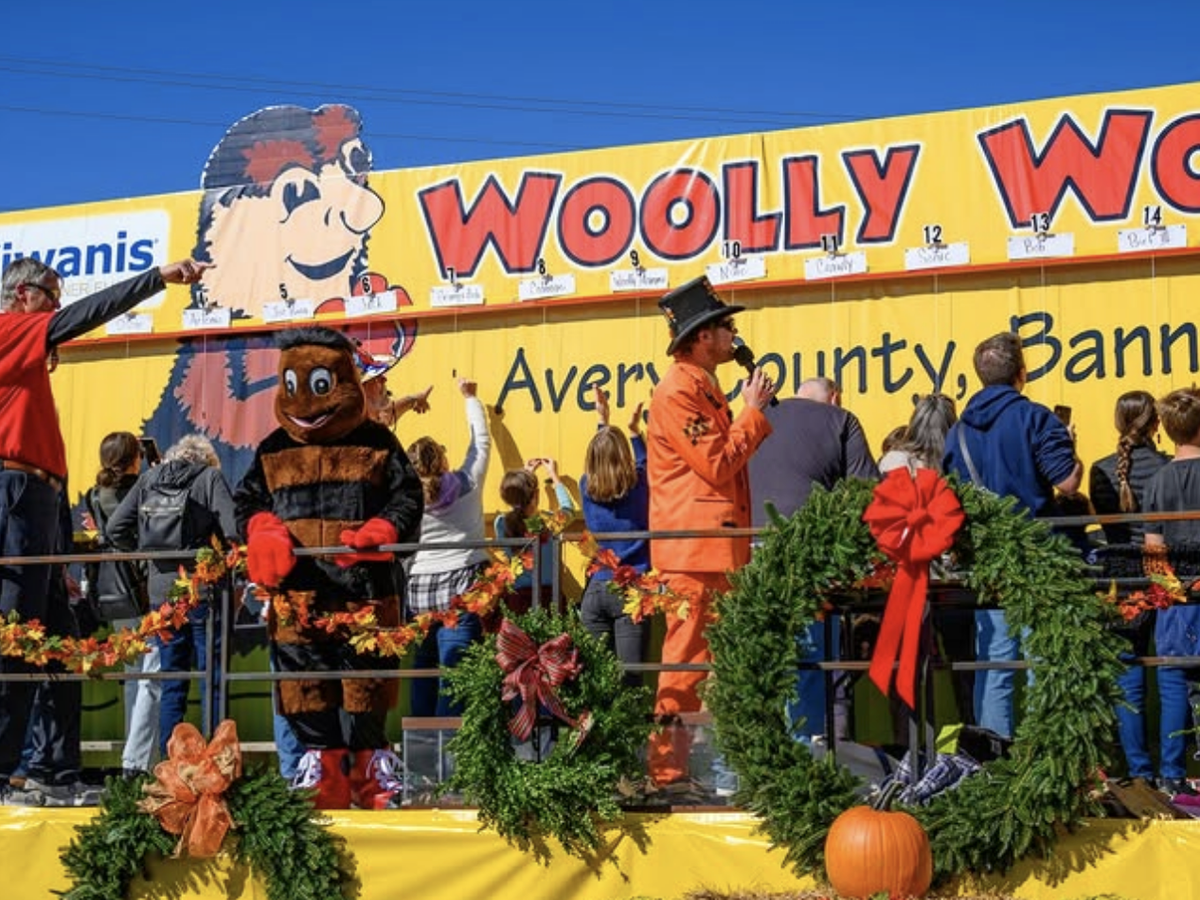 A festive crowd gathers at the Woolly Worm Festival in Banner Elk, North Carolina, featuring a large yellow backdrop with a cartoon worm, a person in a woolly worm mascot costume, a man in an orange suit with a microphone, and children placing worms on strings for a race