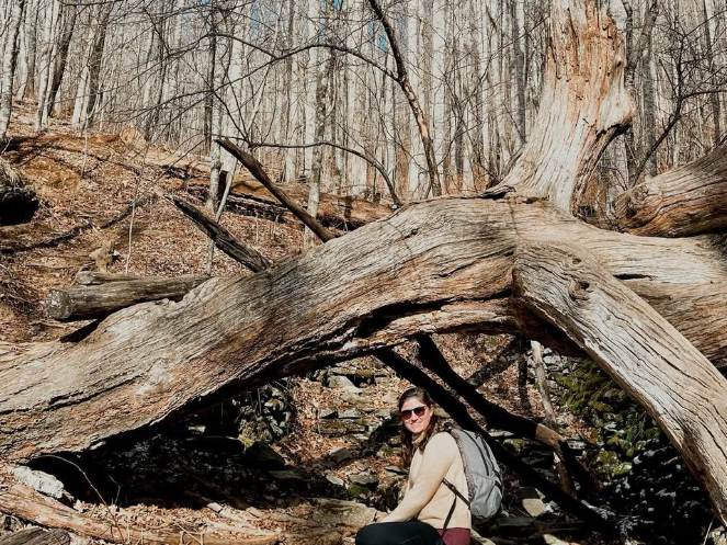 A woman with a backpack crouches and smiles under a massive fallen tree in a leaf-covered forest.