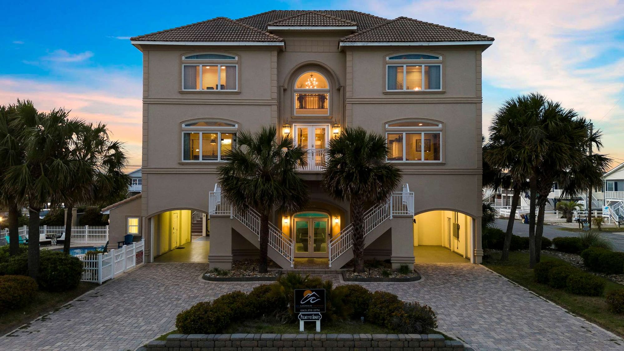 he Palmetto Sunset vacation rental in North Myrtle Beach stands three stories tall with warm lights glowing from every window at dusk, surrounded by tall palm trees and a paved brick driveway. The Mediterranean-style home has a grand dual staircase entry, arched windows, a tiled roof, and a glimpse of a pool tucked to the side — built for big groups who want to be close to the beach.