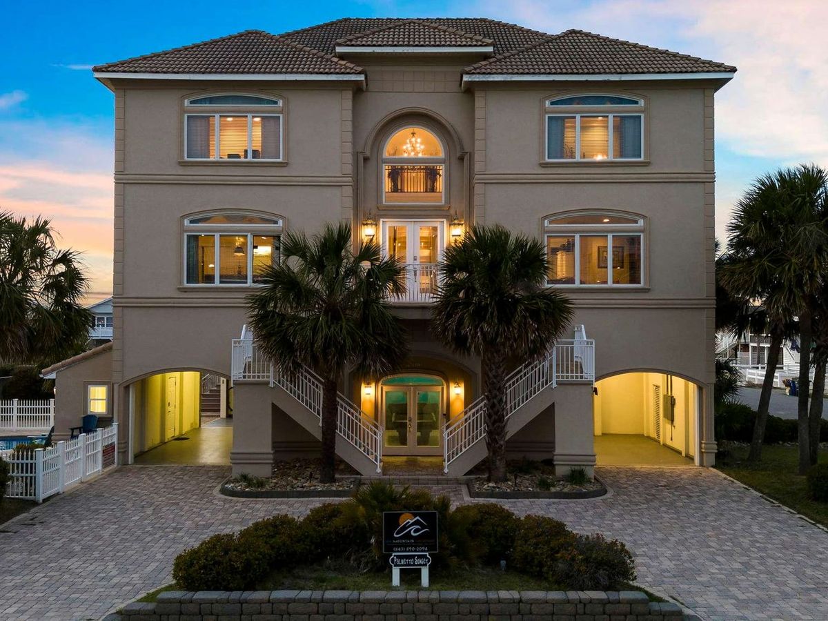 he Palmetto Sunset vacation rental in North Myrtle Beach stands three stories tall with warm lights glowing from every window at dusk, surrounded by tall palm trees and a paved brick driveway. The Mediterranean-style home has a grand dual staircase entry, arched windows, a tiled roof, and a glimpse of a pool tucked to the side — built for big groups who want to be close to the beach.