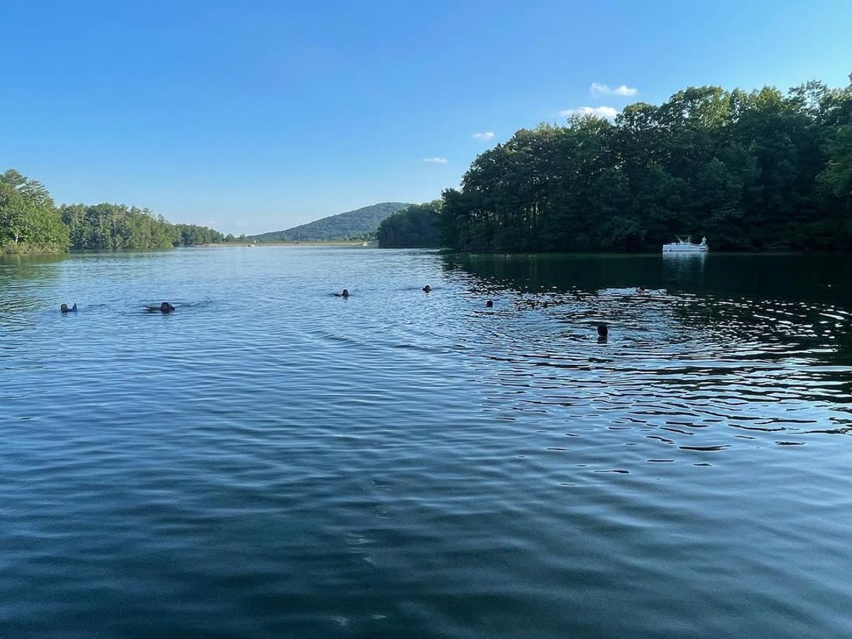 A peaceful summer scene at Lake Petit in Big Canoe, GA, where swimmers enjoy the calm blue water surrounded by lush green trees and distant mountains. A small boat rests along the shoreline, adding to the lake’s relaxing and scenic atmosphere.
