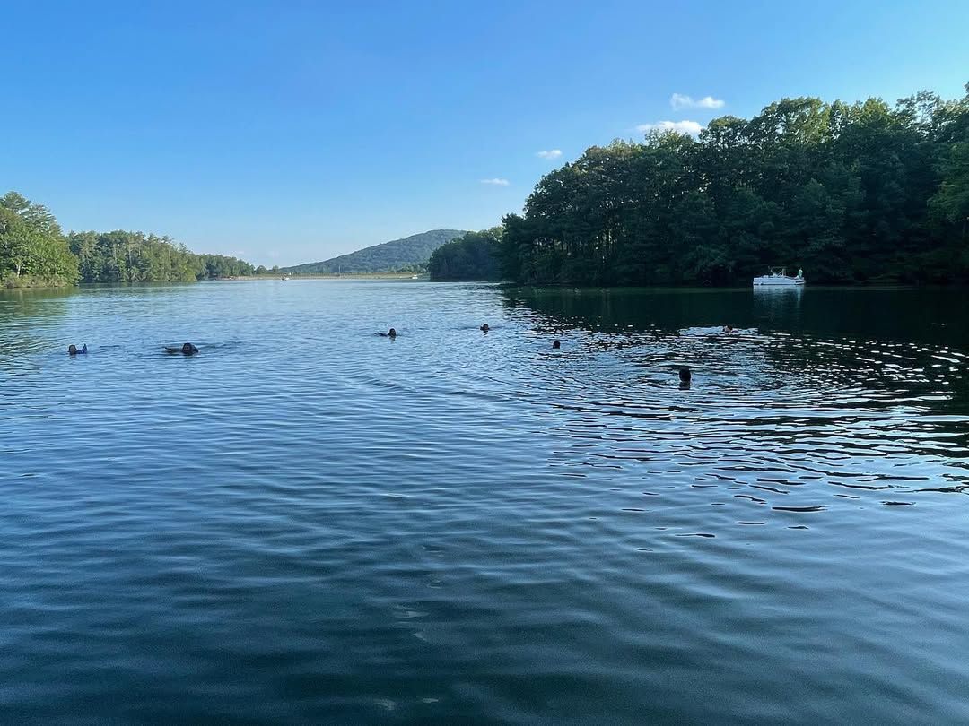 A peaceful summer scene at Lake Petit in Big Canoe, GA, where swimmers enjoy the calm blue water surrounded by lush green trees and distant mountains. A small boat rests along the shoreline, adding to the lake’s relaxing and scenic atmosphere.