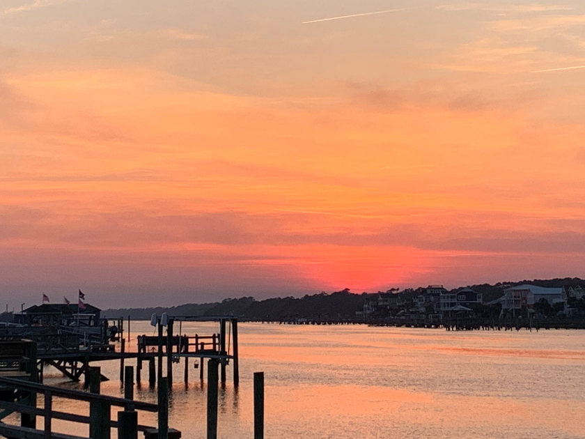 A soft orange and pink sunset lights up the sky over the calm bay, with wooden docks and beach houses lining the water. The perfect quiet end to your Holden Beach day.