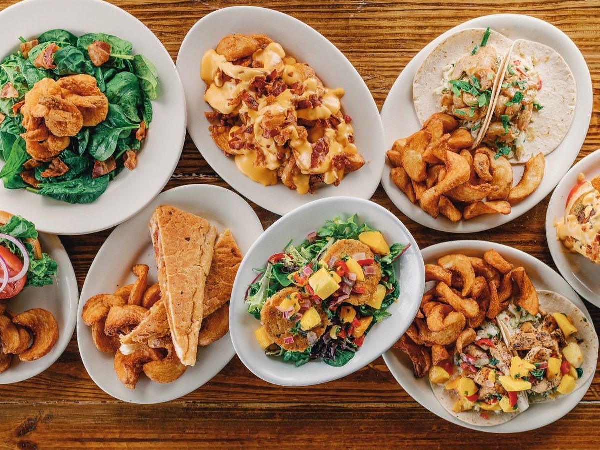 A playful mix of tacos, curly fries, salads, and sandwiches laid out on a wooden table. Southern comfort food meets coastal flair in every dish.