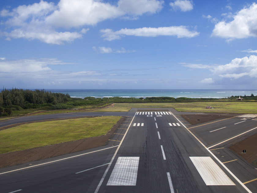 A runway at a small Hawaiian airport stretches toward the ocean, framed by green trees and a partly cloudy sky. A quiet start or end to your island adventure.
