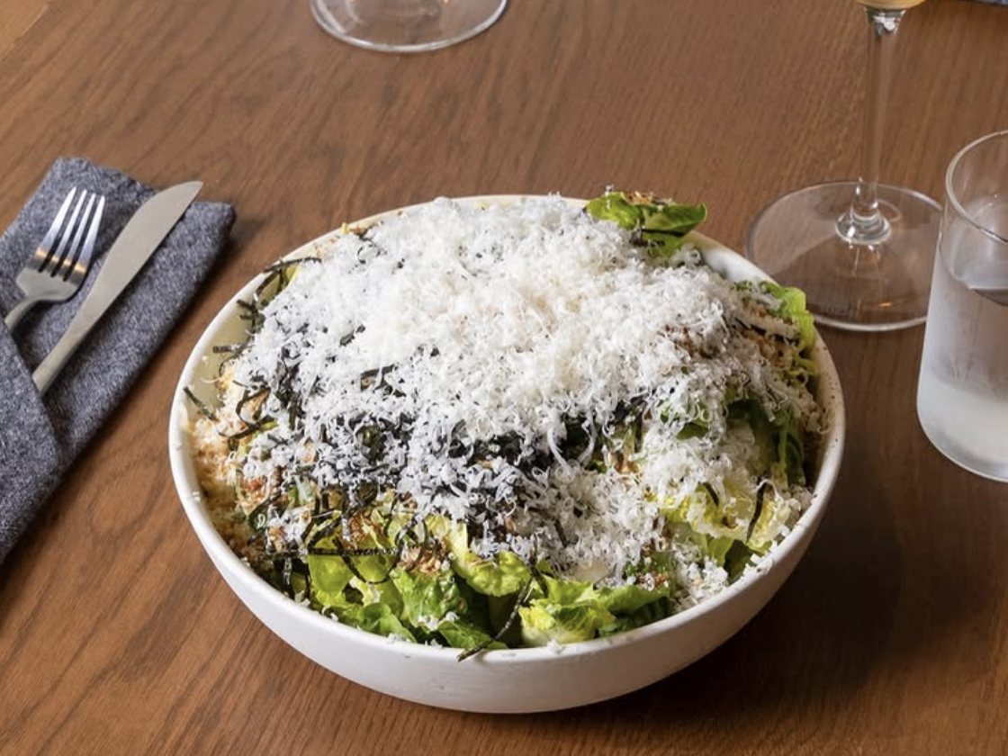 A bowl of fresh green salad generously topped with finely shredded cheese and seaweed, set on a wooden table with a glass of wine, water, and cutlery nearby.
