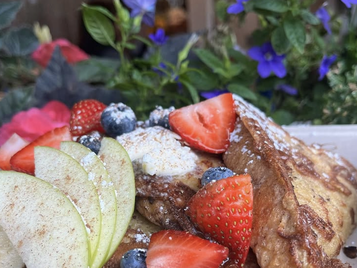 French toast topped with powdered sugar, fresh strawberries, blueberries, sliced green apples, and a dollop of whipped butter, served in a garden setting with colorful flowers in the background.