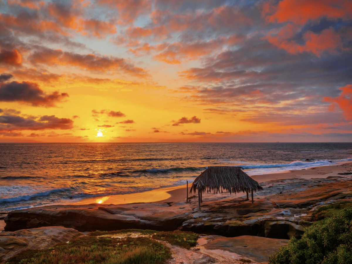 A vibrant sunset over the ocean with waves rolling onto the sandy shore, clouds glowing orange and purple, and a rustic thatched hut standing on the rocky beachfront.