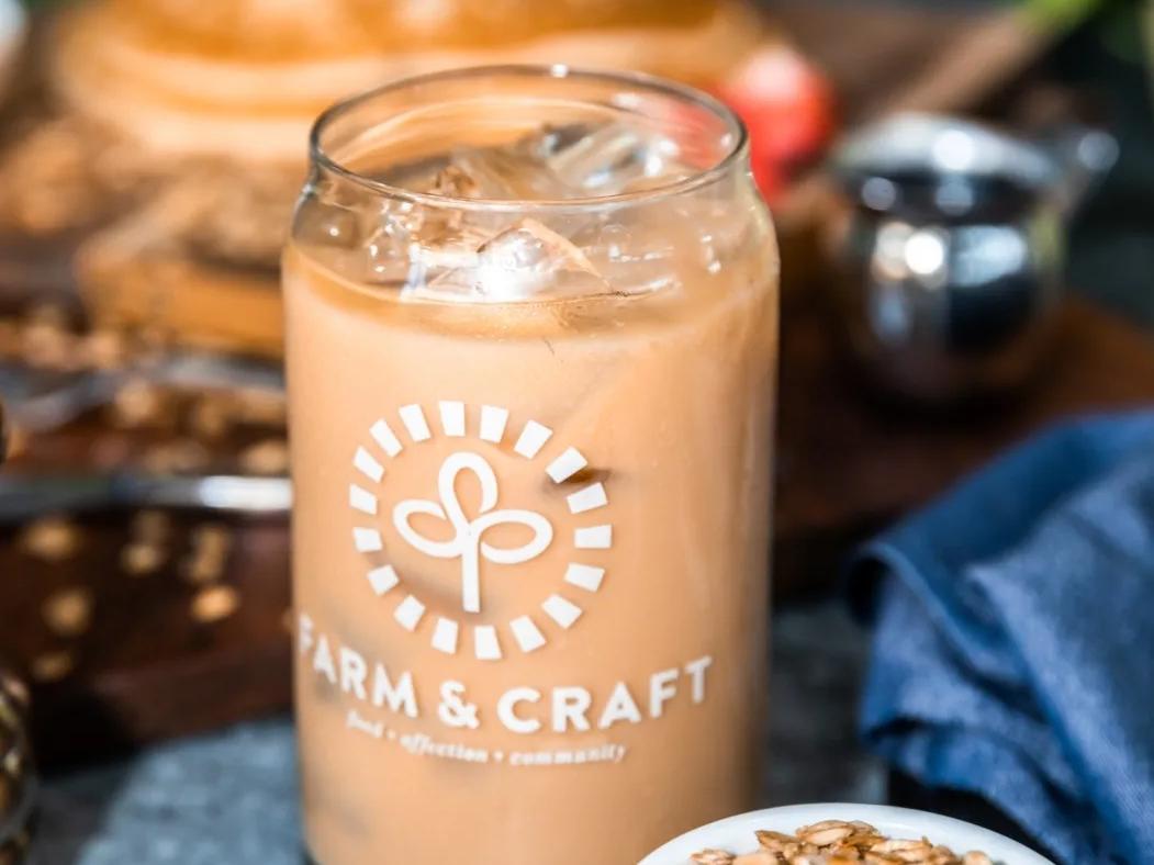 A refreshing iced latte served in a branded glass at Farm & Craft, with fresh granola and a plate of pancakes blurred in the background. The café highlights healthy, sustainable dining with a modern touch.