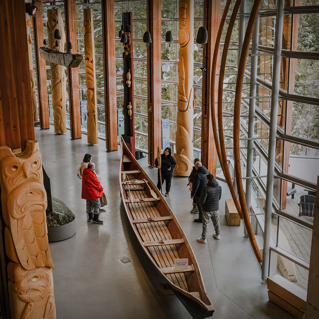 Visitors explore carved totem poles and a traditional canoe inside the Squamish Lil’wat Cultural Centre. Large windows and natural wood design highlight Indigenous culture and history in Whistler. This cultural center shares the stories of the Squamish and Lil’wat Nations.
