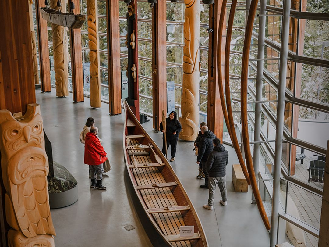 Visitors explore carved totem poles and a traditional canoe inside the Squamish Lil’wat Cultural Centre. Large windows and natural wood design highlight Indigenous culture and history in Whistler. This cultural center shares the stories of the Squamish and Lil’wat Nations.