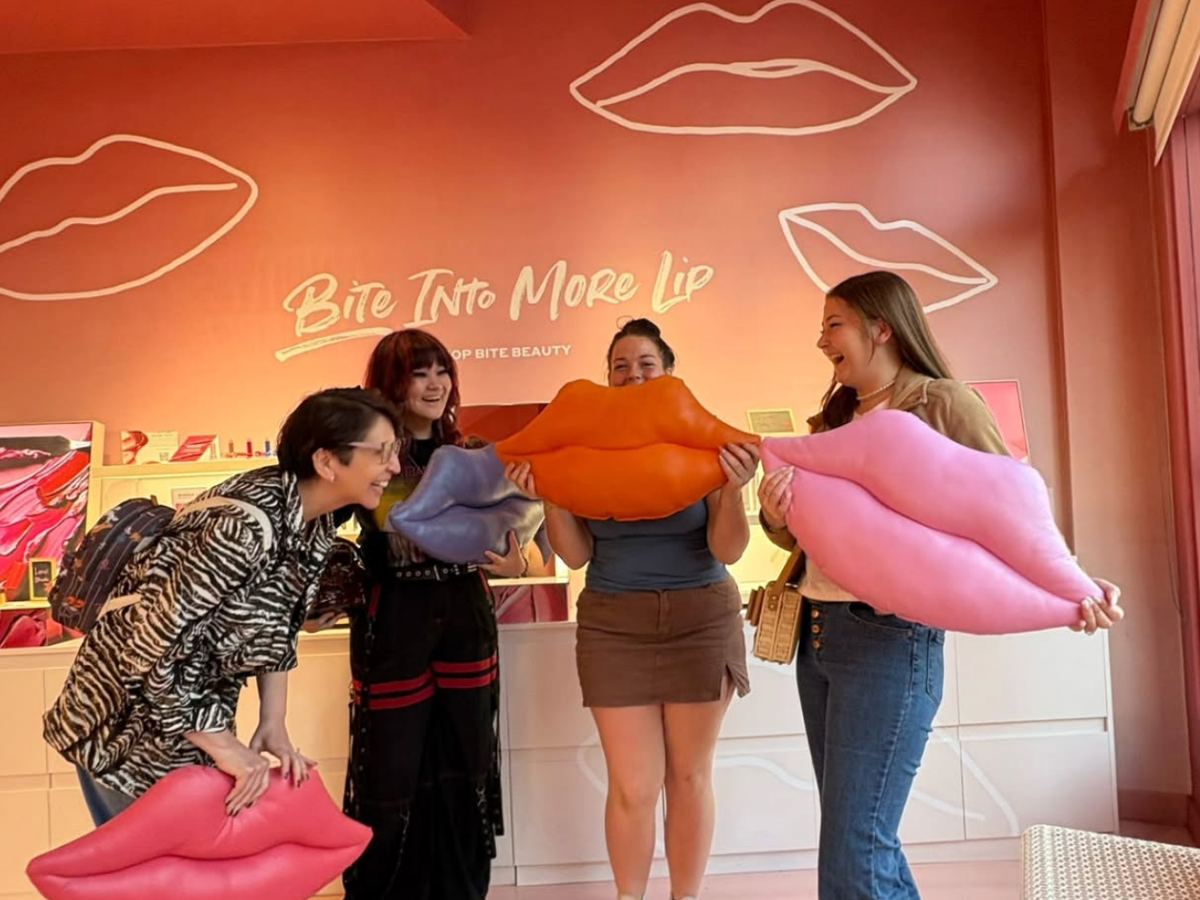 Four women laugh and pose with oversized lip-shaped pillows in a vibrant lipstick-themed store with a neon sign that reads "Bite Into More Lip."
