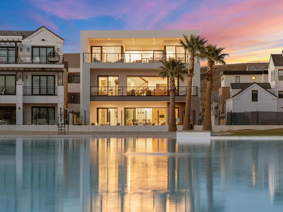 A modern three-story luxury home with large glass windows and balconies, overlooking a serene lagoon at sunset. The house features contemporary architecture with clean lines, warm interior lighting, and palm trees in the foreground, reflecting on the calm water