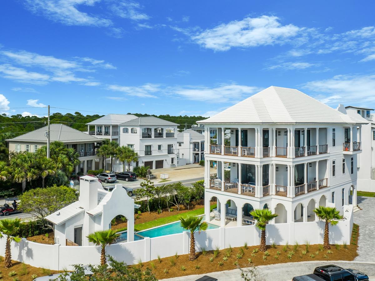 A grand three-story white coastal home with wraparound balconies sits behind a private white fence enclosing a pool and outdoor fireplace, surrounded by palm trees and neighboring beach houses.
