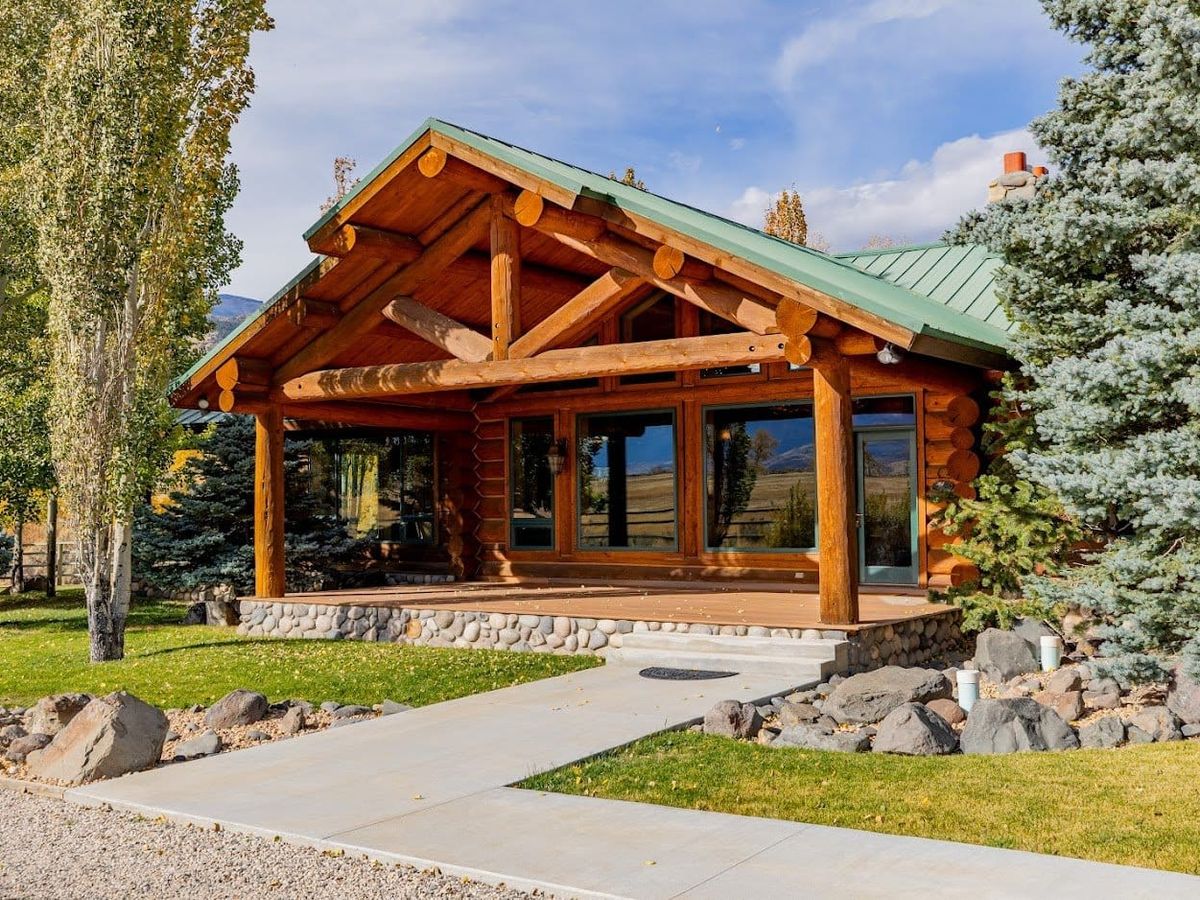 A rustic log cabin with a green roof sits among trees and open land in Cody, Wyoming. This cozy home base offers easy access to Yellowstone and the surrounding Western adventures.