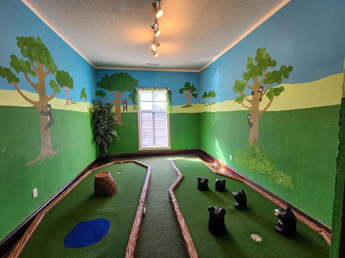 A colorful indoor mini golf course with hand-painted murals of trees and bears creates a playful atmosphere. The narrow green paths and fun obstacles make it a perfect family activity. Sunlight streams through a window, brightening the whimsical space.