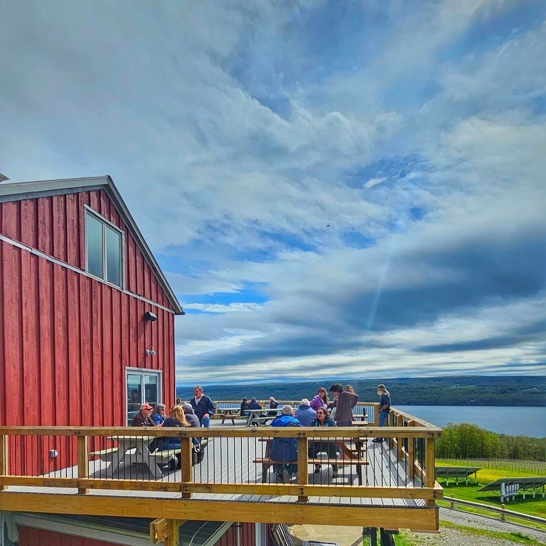 Visitors sit on the deck at Two Goats Brewing, overlooking Seneca Lake on a clear day. The casual outdoor space is perfect for enjoying a cold beer with lake views.