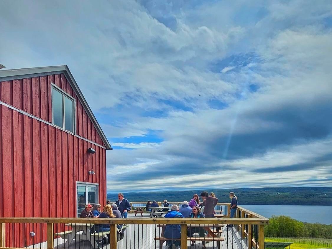 Visitors sit on the deck at Two Goats Brewing, overlooking Seneca Lake on a clear day. The casual outdoor space is perfect for enjoying a cold beer with lake views.
