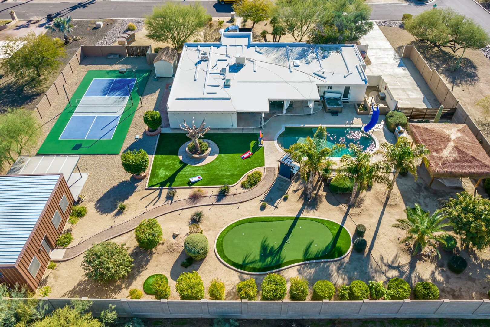 Aerial view of a desert estate featuring a pickleball court, putting green, pool with a slide, and lush landscaping.