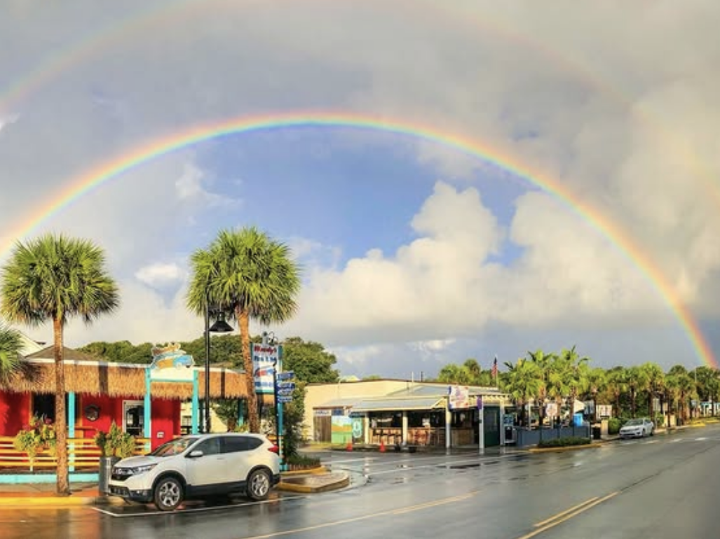 A vibrant double rainbow arcs across a coastal town street lined with palm trees, colorful buildings, and wet pavement after a rain shower.
