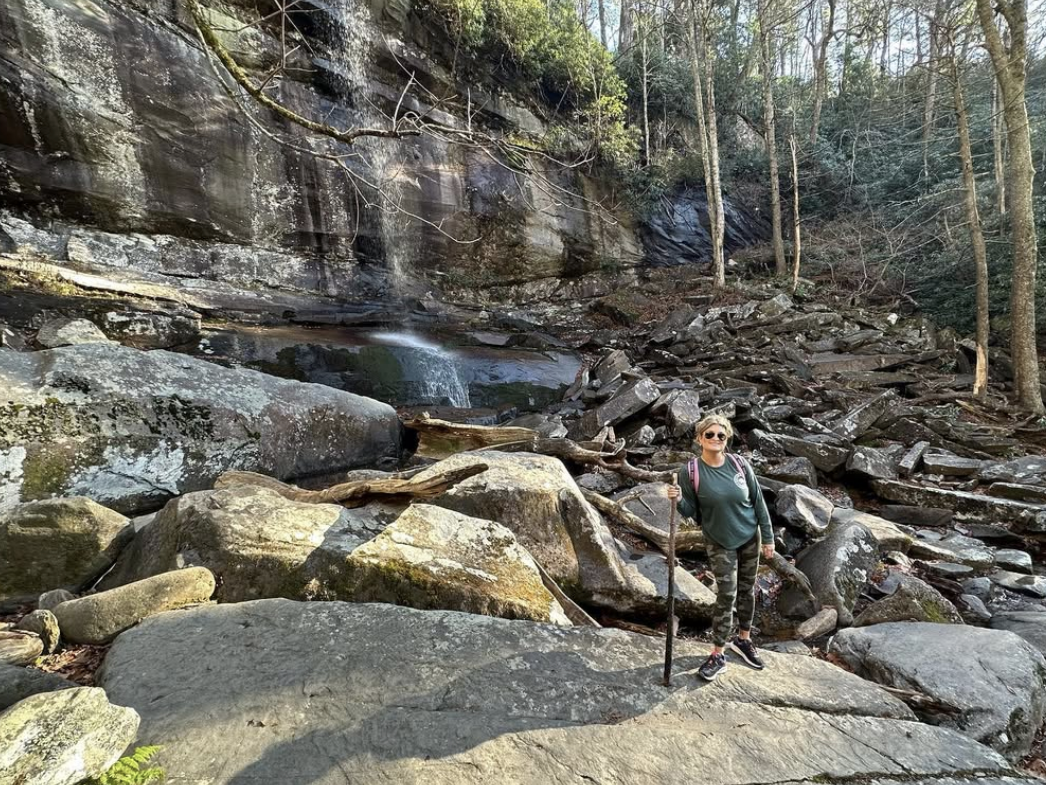 A woman with a hiking stick poses confidently on large boulders near a gentle waterfall and rocky cliffside in a forested area of the Great Smoky Mountains.