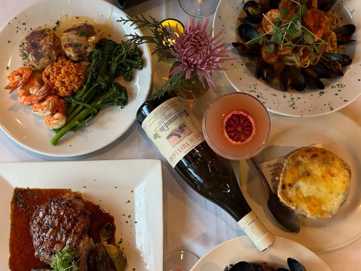 Array of food on white plates and bowls with flower vase, drinks, and wine bottle displayed on table