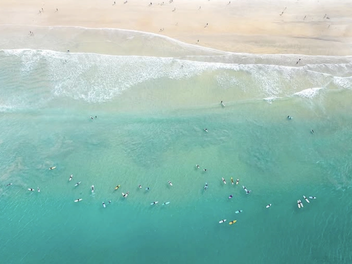 Aerial view of surfers floating on their boards in turquoise water near the shore, with gentle waves breaking and people walking along the sandy beach.