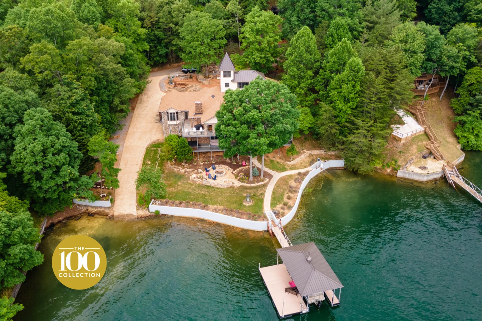 Aerial view of a luxury lakefront estate surrounded by dense green forest, featuring a private dock, winding shoreline walkway, fire pit seating area, and multiple waterfront structures on calm emerald water.