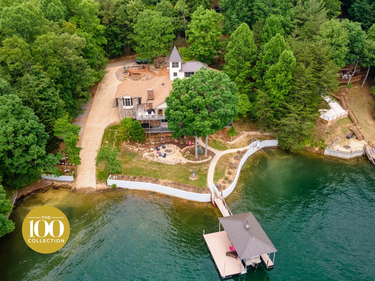 Aerial view of a luxury lakefront estate surrounded by dense green forest, featuring a private dock, winding shoreline walkway, fire pit seating area, and multiple waterfront structures on calm emerald water.