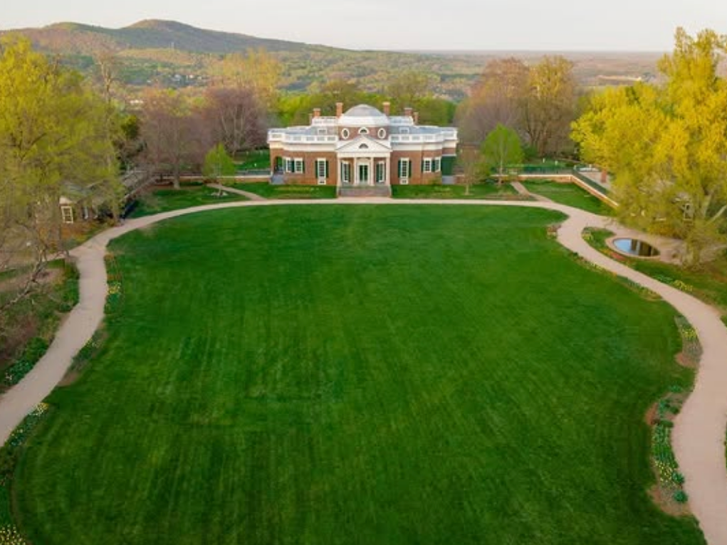 A stately historic mansion with a white dome and columns sits at the edge of a large, manicured lawn, framed by curved walking paths and surrounded by trees and distant hills.