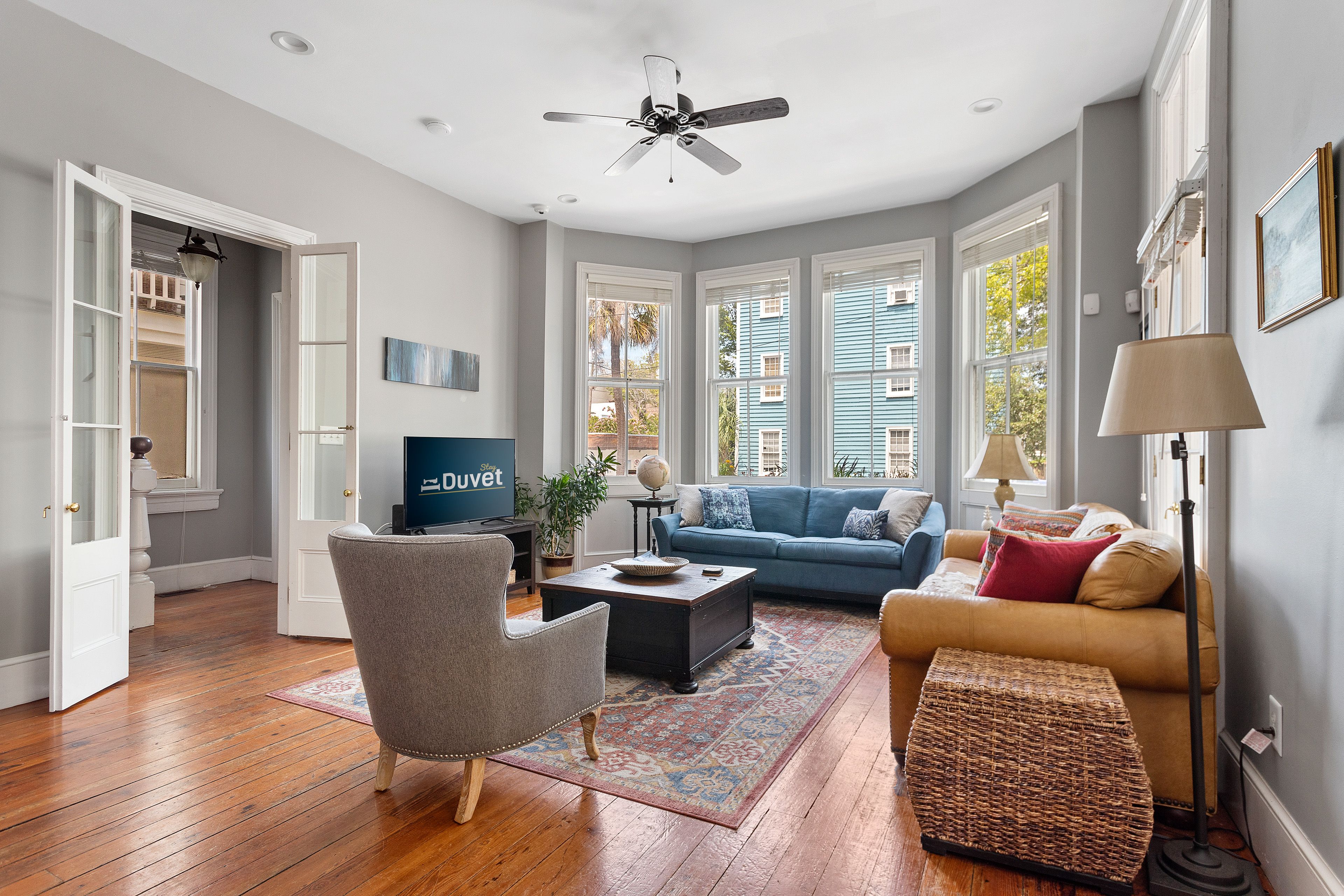 A bright and inviting living room features a mix of cozy seating including a blue couch, tan loveseat, and gray armchair, with large windows letting in natural light and views of colorful buildings outside.