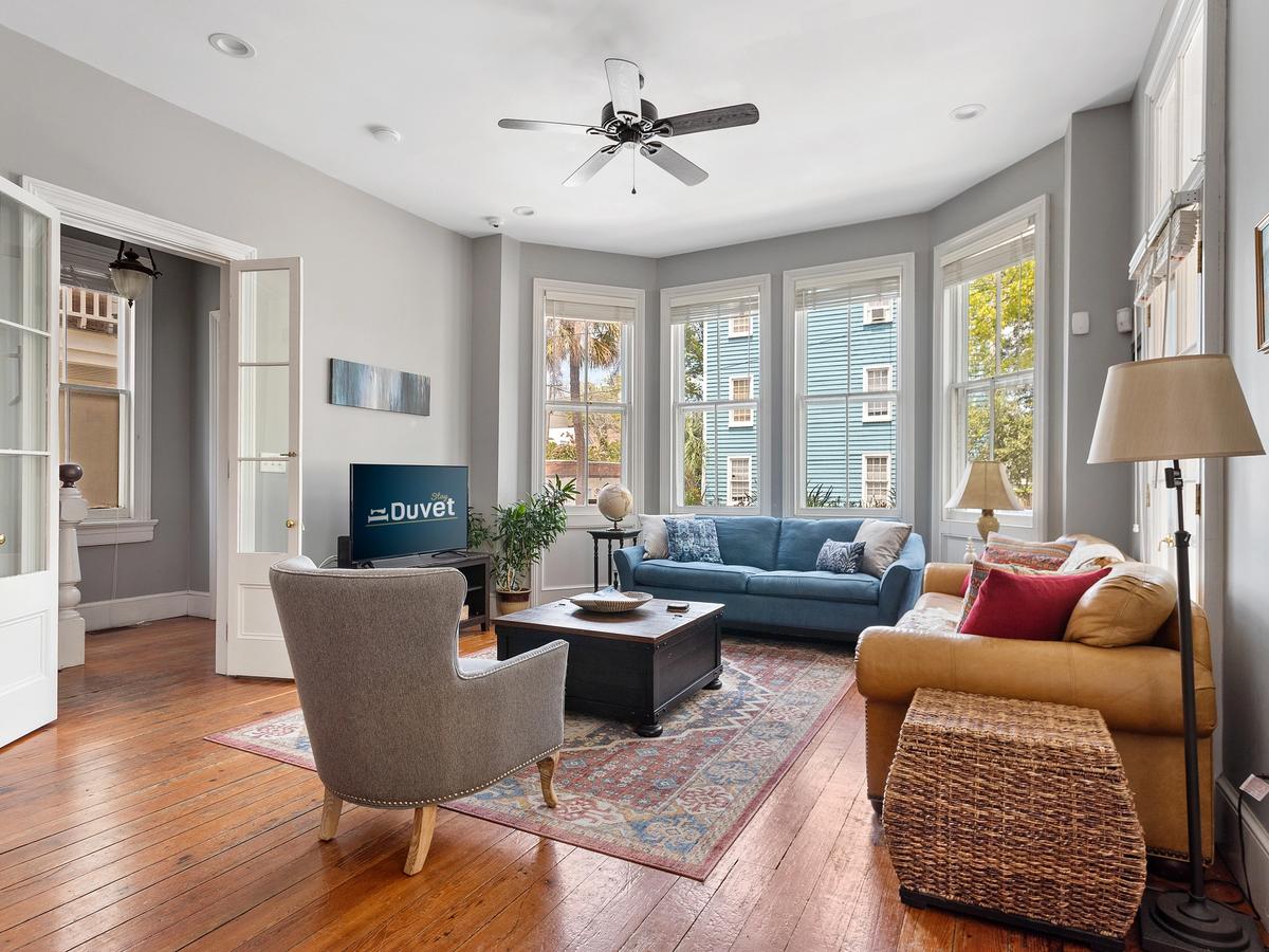 A bright and inviting living room features a mix of cozy seating including a blue couch, tan loveseat, and gray armchair, with large windows letting in natural light and views of colorful buildings outside.