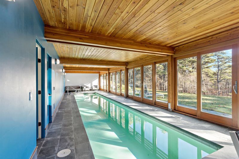 Indoor lap pool with a vaulted wood-paneled ceiling, floor-to-ceiling windows overlooking a wooded yard, slate tile surround, and lounge seating at the far end for a serene, year-round retreat.