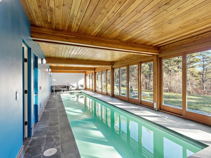 Indoor lap pool with a vaulted wood-paneled ceiling, floor-to-ceiling windows overlooking a wooded yard, slate tile surround, and lounge seating at the far end for a serene, year-round retreat.
