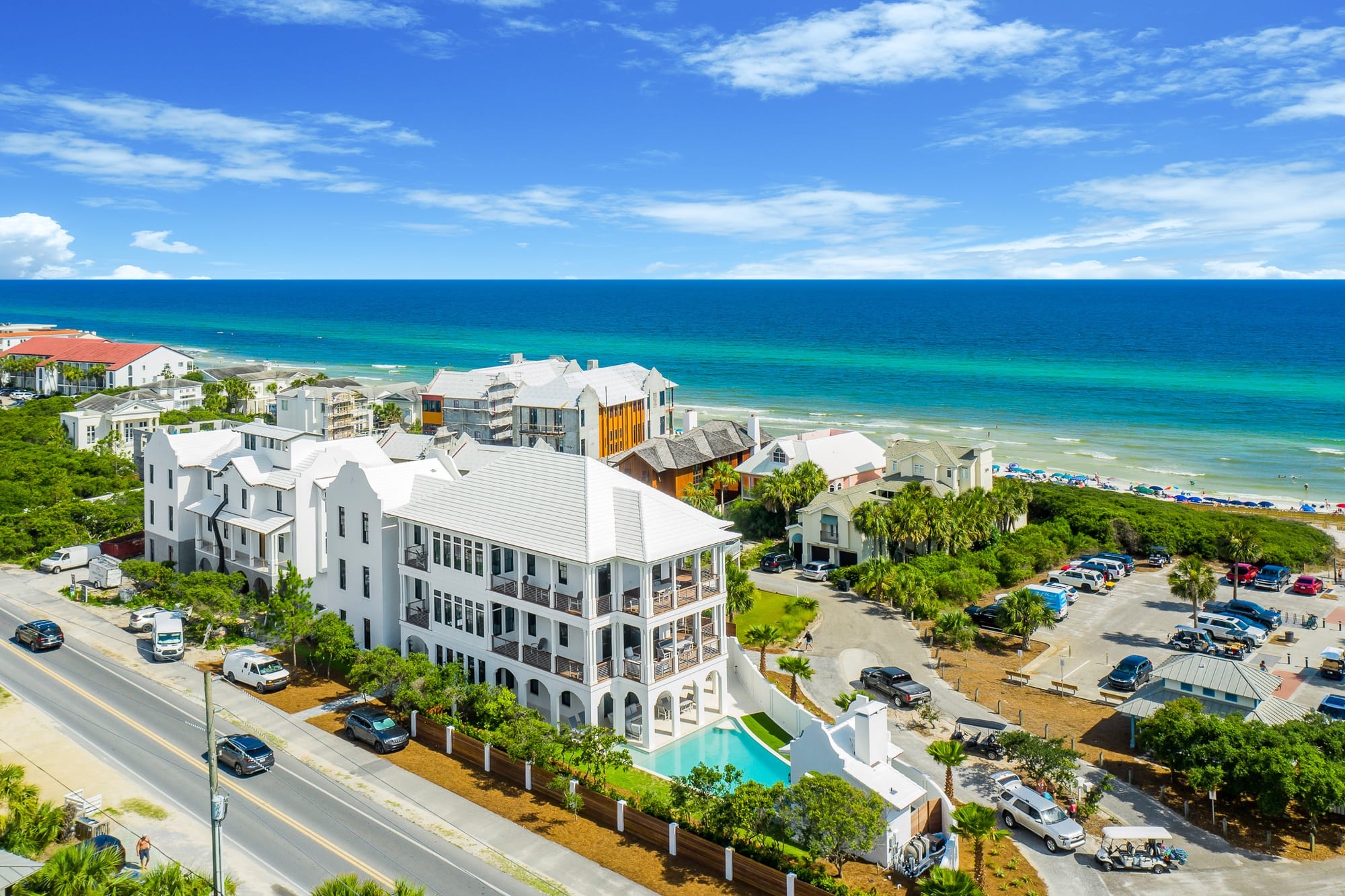 A luxury white beach estate overlooks bright turquoise water along a sunny coastline. The clean, modern architecture and private pool make the home stand out among the other vacation houses nearby. The view captures a lively beach scene, perfect for travelers who love sun, sea, and stylish coastal living.