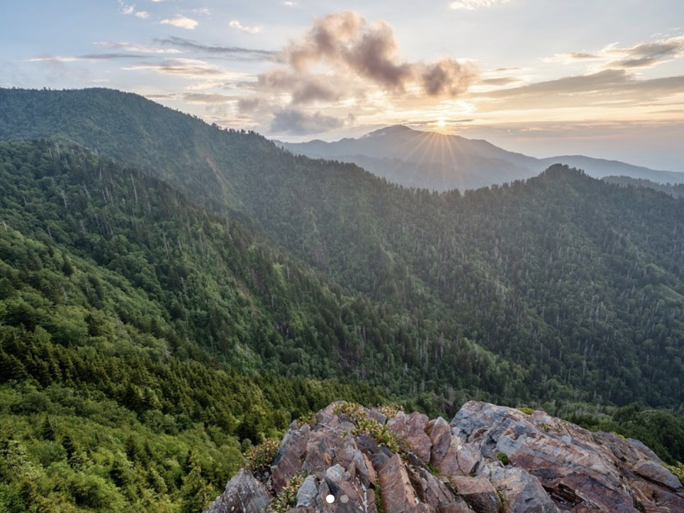 Sunrise over a lush, forested mountain range in the Great Smoky Mountains, viewed from a rocky outcrop with layers of green ridges stretching into the distance.