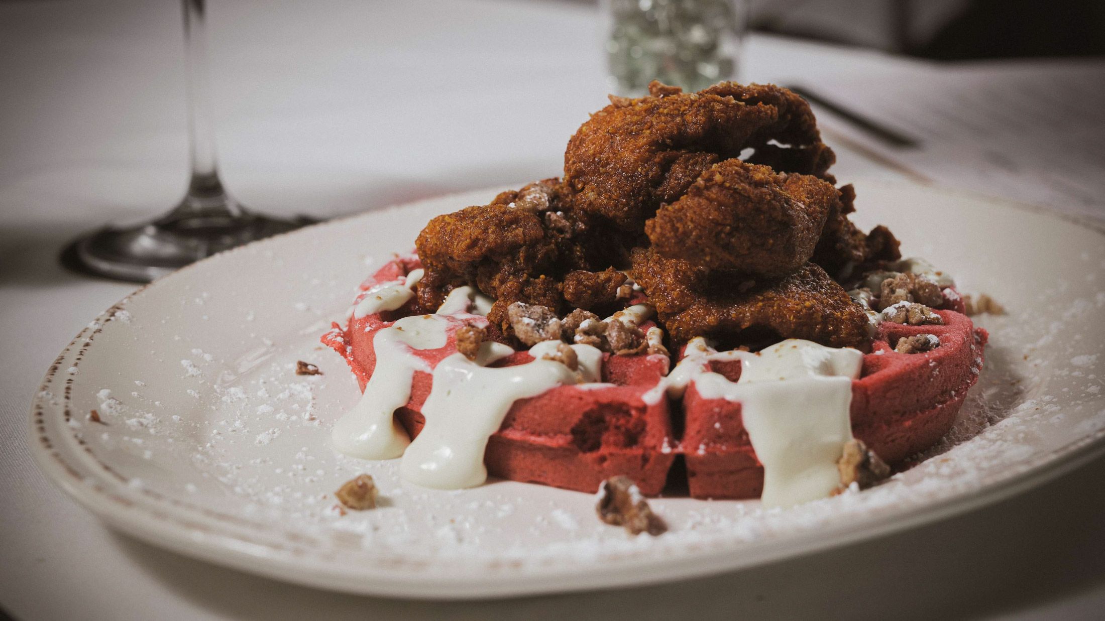 A generous portion of crispy fried chicken sits atop a vibrant red velvet waffle, drizzled with cream sauce and scattered with candied pecans on a white ceramic plate. Powdered sugar dusts the plate and a wine glass is softly visible in the background, hinting at an upscale dining setting