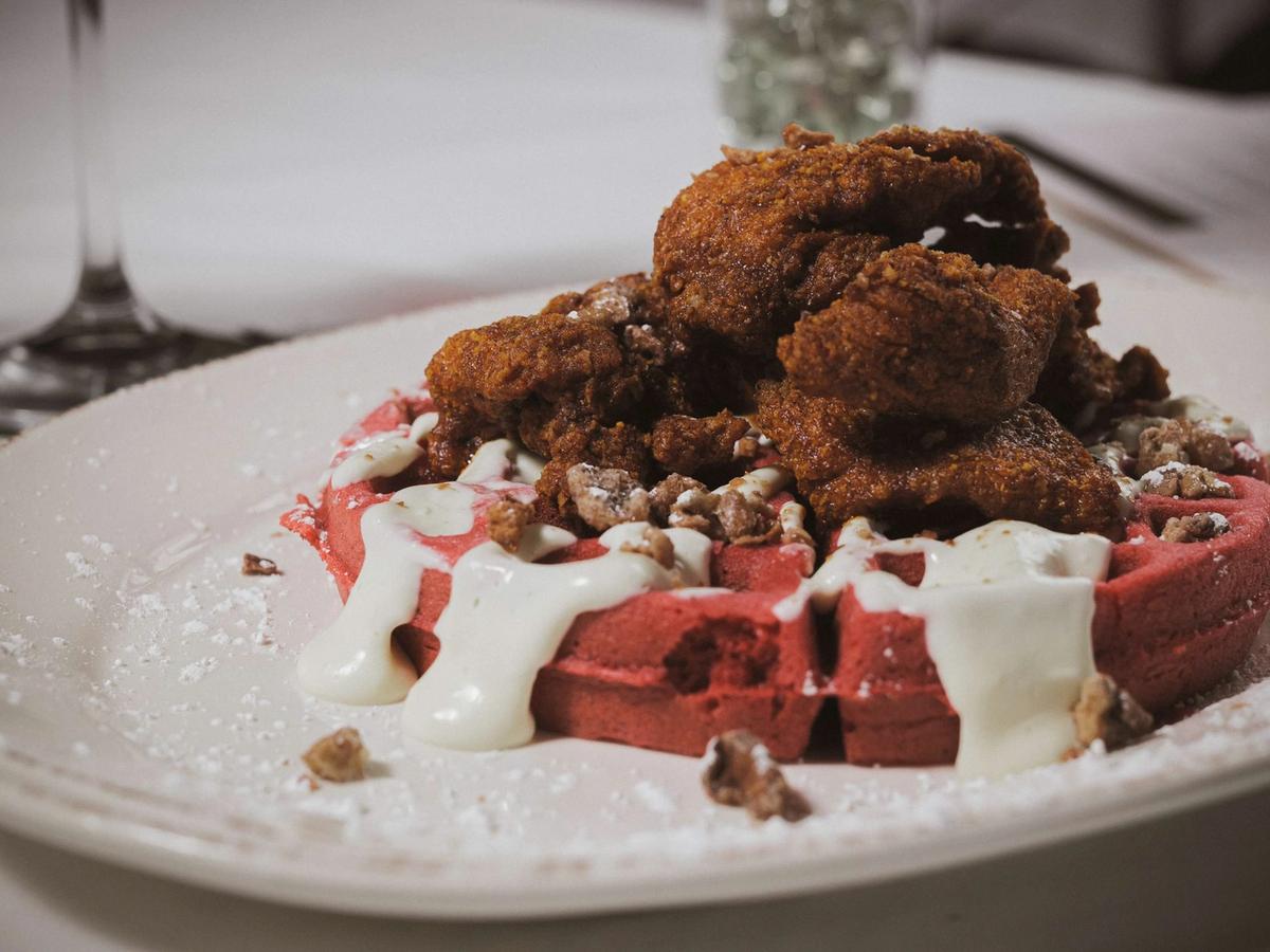 A generous portion of crispy fried chicken sits atop a vibrant red velvet waffle, drizzled with cream sauce and scattered with candied pecans on a white ceramic plate. Powdered sugar dusts the plate and a wine glass is softly visible in the background, hinting at an upscale dining setting