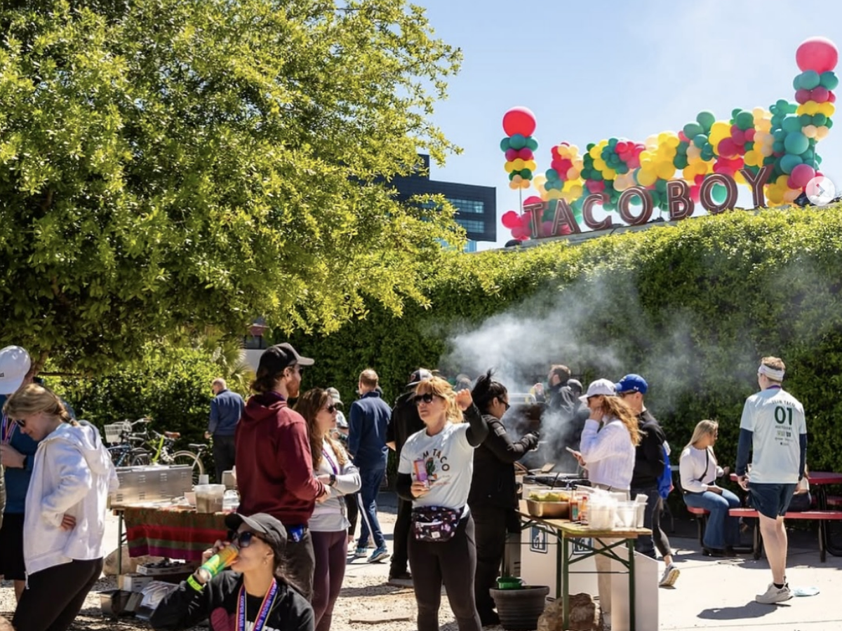 A lively outdoor gathering in front of Taco Boy, with people mingling, drinking, and cooking under a sunny sky and colorful balloon display.